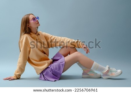 Fashionable confident girl wearing purple sunglasses, pleated mini skirt, yellow hoodie, socks, high top colorful sneakers, sitting, posing on blue background. Full-length studio portrait. Copy space Royalty-Free Stock Photo #2256706961