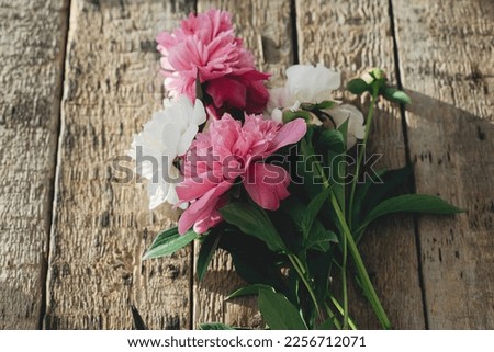 Beautiful peony bouquet in sunlight on rustic aged wood. Stylish floral greeting card. Fresh pink and white peony flowers on wooden table, moody image. Gathering flowers in countryside
