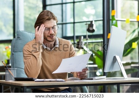 A young blond businessman is dissatisfied with the result of financial work with documents, the financier looks at the reports and contract the man is inside the office on paper work with computer. Royalty-Free Stock Photo #2256715023