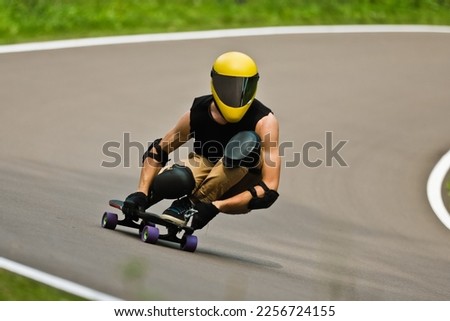 A cool man in a yellow helmet and leather suit, in a rack at high speed, rides on a long longboard for downhill on asphalt
