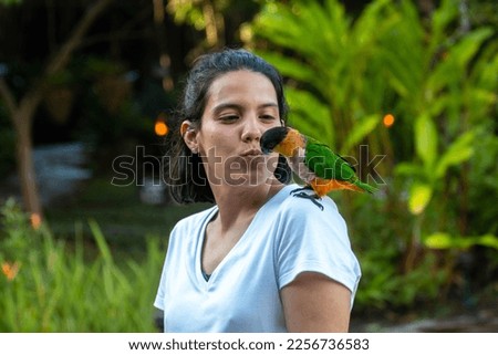 A girl kisses a green parrot that sits on her shoulder in a garden