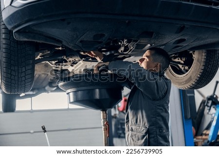 A car workshop worker is draining oil from engine while standing under the car. Royalty-Free Stock Photo #2256739905