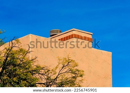 Block style chimney on an adobe house with tiny red tiles or bricks on vent with metal top and front or back yard tree. In late afternoon sun in the southwestern neighborhood on home in neighborhood.