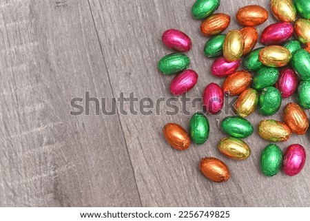 Foil wrapped multi coloured easter eggs in pink, green, orange and yelow in a pile or group, against a grey white wooden background. 