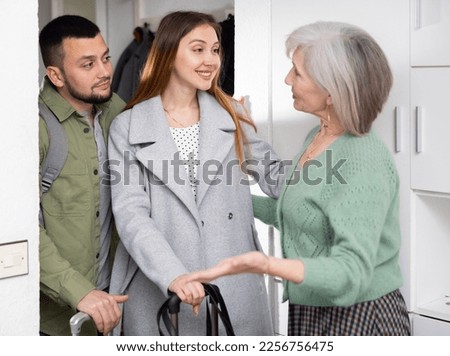 Positive elderly woman meeting young couple at doorway and making welcoming gesture Royalty-Free Stock Photo #2256756475