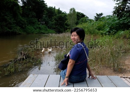 An Asian female tourist sitting on a wooden deck enjoying view of a muddy river and a group of duck in Kanchanaburi province in Thailand.