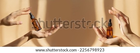 Two pairs of female hands hold amber glass vials with cosmetic serum on light brown background, in rays of sunlight. Close-up, banner. Branding, mockup concept. Royalty-Free Stock Photo #2256798923