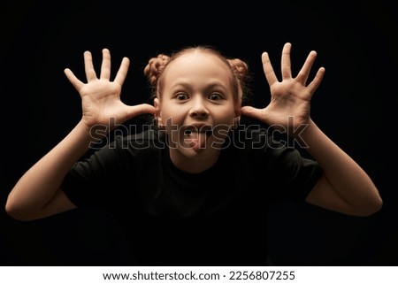Portrait of a cute funny girl in a black t-shirt making faces on a black studio background. Children, emotions. Psychological picture.
