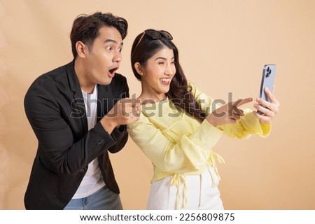 Shocked excited Asian businessman along with happy smiling woman pointing finger at smart phone over beige studio background. Man in white shirt and suit and woman in yellow top and white trousers. Royalty-Free Stock Photo #2256809875