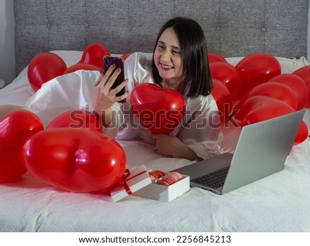 Woman in white communicates online with computer while lying on a bed with red heart shape balloons and gift box with macarons. Woman works at home, using laptop. Valentines Day, wedding. Copy space.
