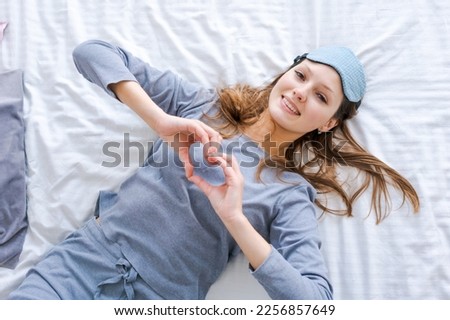 Happy healthy, beautiful young woman wearing sleep mask and blue pajamas, awake after sleeping, laughing lies in comfortable bed on pillow, smiling lady wakes up, enjoy good morning