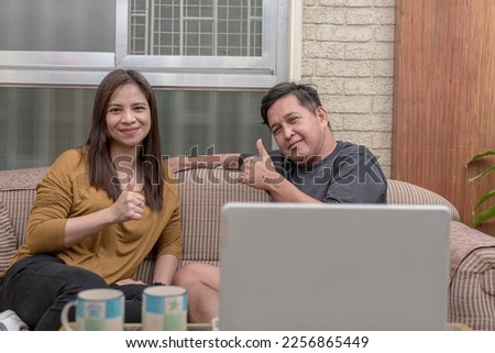 A happy asian couple in their 40s making a thumbs up sign of approval while relaxing on the sofa at the living room.