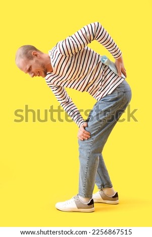 Young man warming his lower back with hot water bottle on yellow background