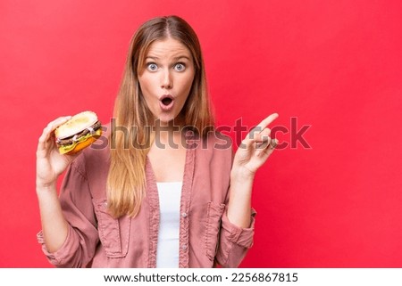 Young caucasian woman holding a burger  isolated on red background surprised and pointing side