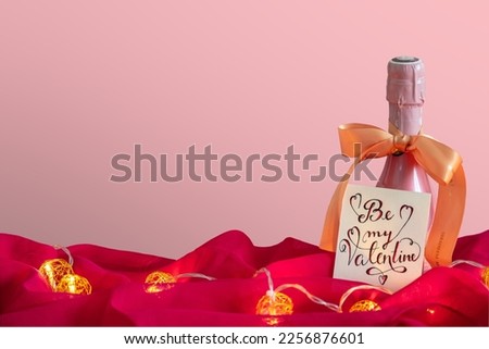Romantic Valentine background with copy space isolated champagne bottle with golden ribbon bow and love handwritten calligraphic note Be my valentine, pink satin and light chain for romantic dinner