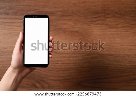 Woman holding smartphone with blank screen at wooden table, top view. Mockup for design