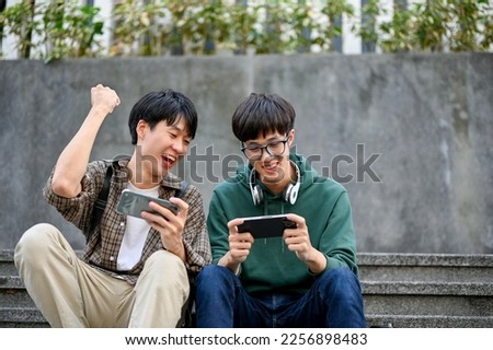 Cheerful and overjoyed young Asian male college student playing mobile game with his friend, cheering his friend, having a fun time together.