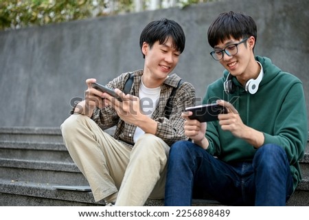 Happy young Asian male college student playing mobile game with his friend, cheering his friend, having a fun time together. best friend concept