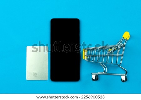 Card, mobile phone with black screen, shopping cart on blue background, copy space, top view. Online shopping and payment concept