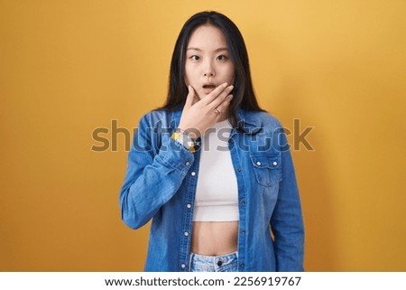 Young asian woman standing over yellow background looking fascinated with disbelief, surprise and amazed expression with hands on chin 