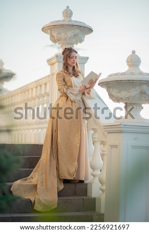 A beautiful young woman in an eighteenth century historical gold dress stands on the stairs of a mansion with a book in her hands. the princess is reading in the palace. fabulous image Royalty-Free Stock Photo #2256921697