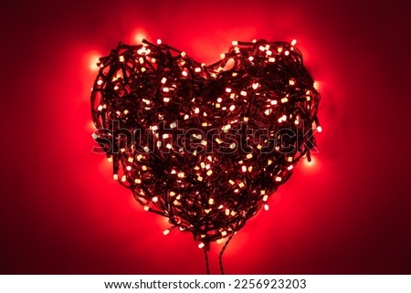 Shape of heart folded from tangled wires of a garland with red light bulbs on red background in the dark with a vignetting effect
