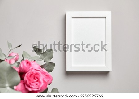 White photo frame mockup hanging on grey concrete wall and and bouquet of pink roses with eucalyptus leaves. Empty blank photo frame and floral decor