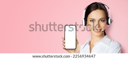 Customer support phone operator in headset holding showing smartphone cell phone mobile white blank mock up screen, isolated rose pink background. Consulting and assistance service call center ad.
