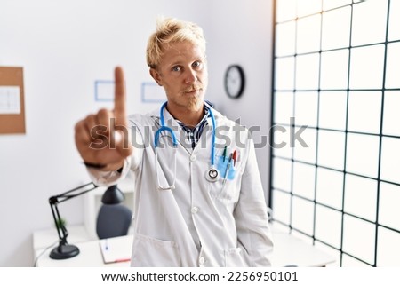 Young blond man wearing doctor uniform and stethoscope at clinic pointing with finger up and angry expression, showing no gesture 