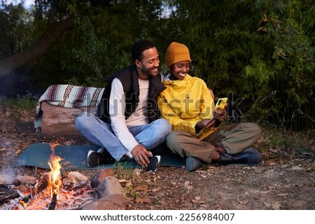 Copy space photo of lovely African American young couple sitting at camping in the mountain with campfire, using mobile phone taking selfies and looking at them happily.