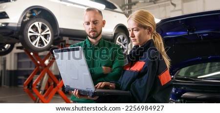 A young female car mechanic holds a diagnostic laptop and shows the manager the identified errors against the background of the car. Panoramic photography