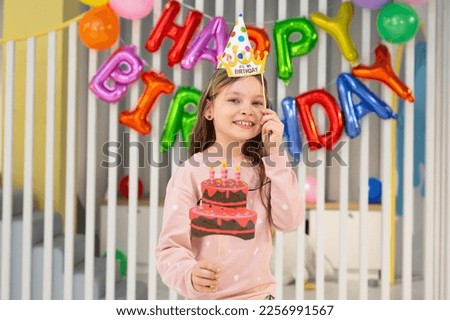 The birthday girl in a festive hat, with a pipe and props is having fun on the background of a multi-colored wall with balloons