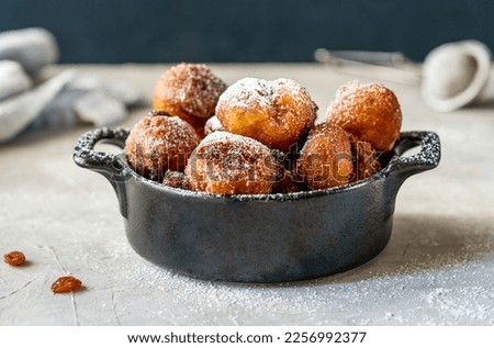 Italian traditional carnival fritters made with raisins and topped with sugar powder set in black pot on concrete table, strainer, napkin, raisins. Dark background. Close up Royalty-Free Stock Photo #2256992377