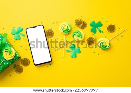 St Patrick's Day concept. Top view photo of smartphone shamrocks meringue lollipops sprinkles gold coins and giftbox on isolated vibrant yellow background with copyspace