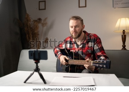Student learning to play guitar at home or online tutor. Smiling man looks at guitar and makes video lesson for followers in living room. Copy space
