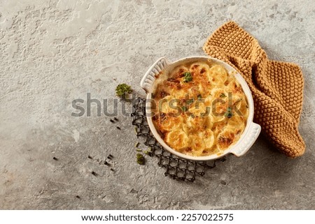 Top view of palatable scalloped gratin potato with herbs and metal stand for hot dish near towel on gray stone surface Royalty-Free Stock Photo #2257022575