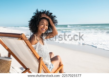 Portrait of happy african american woman sunbathing on wooden deck chair at tropical beach while looking at camera. Smiling black girl enjoying vacation at seaside with copy space. Woman relaxing. Royalty-Free Stock Photo #2257025577