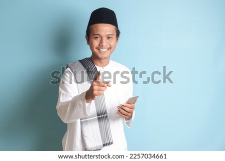 Portrait of young Asian muslim man holding mobile phone with smiling expression on face while pointing finger to the side. Isolated image on blue background