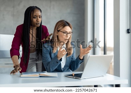 Two multiethnic business women in suit having problems with laptop, system being hacked stopped working lost internet connection in middle of important work, feeling frustrated over deleted lost data. Royalty-Free Stock Photo #2257042671