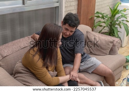 A supportive middle aged man comforts his depressed and disappointed wife, holding her hand while sitting together at the couch in the living room. Royalty-Free Stock Photo #2257045537