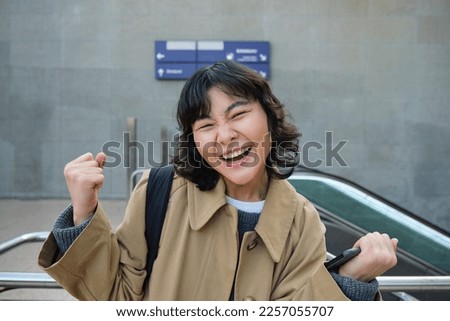 Enthusiastic asian girl stands on street, makes hooray celebration gesture, says yes, triumphing, winning and enjoying this feeling, standing alone outdoors.