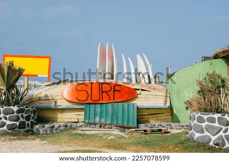 The red surfboard with the inscription Surf hanging on a old wooden boat surrounded by billboard and palm trees against blue sky in sunlight. Copy space. Advertisement for surfing school.