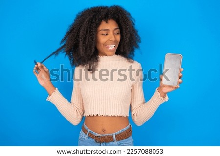 Photo of nice pretty Young woman with afro hair style wearing crop top over blue background demonstrate phone screen hold hair tails