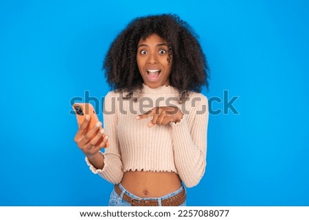 Portrait cute astonished Young woman with afro hair style wearing crop top over blue background impressed unbelievable unexpected incredible notification advice