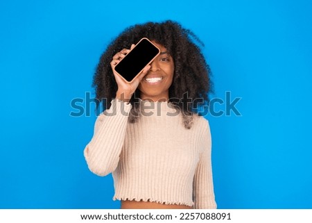 Young woman with afro hair style wearing crop top over blue background holding modern smartphone covering one eye while smiling