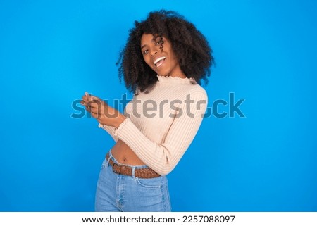 Nice addicted cheerful Young woman with afro hair style wearing crop top over blue background using gadget playing network game