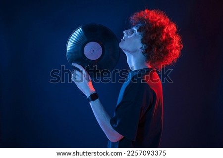Musician holding vinyl. Young man with curly hair is indoors illuminated by neon lighting. Royalty-Free Stock Photo #2257093375