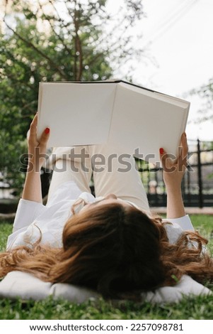 Magazine or book image mockup. A young brunette girl reads a book lying on the lawn in the summer garden.
