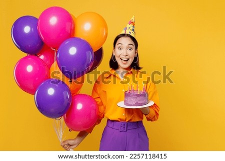 Happy fun excited amazed young woman wears casual clothes hat celebrating hold bunch of colorful air balloons cake with candles isolated on plain yellow background. Birthday 8 14 holiday party concept