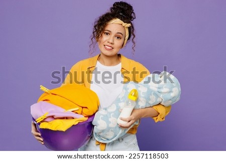 Young sad housekeeper woman wear yellow shirt tidy up hold newborn baby basin with clothes for laundry do everyday routine isolated on plain pastel light purple background studio. Housework concept Royalty-Free Stock Photo #2257118503